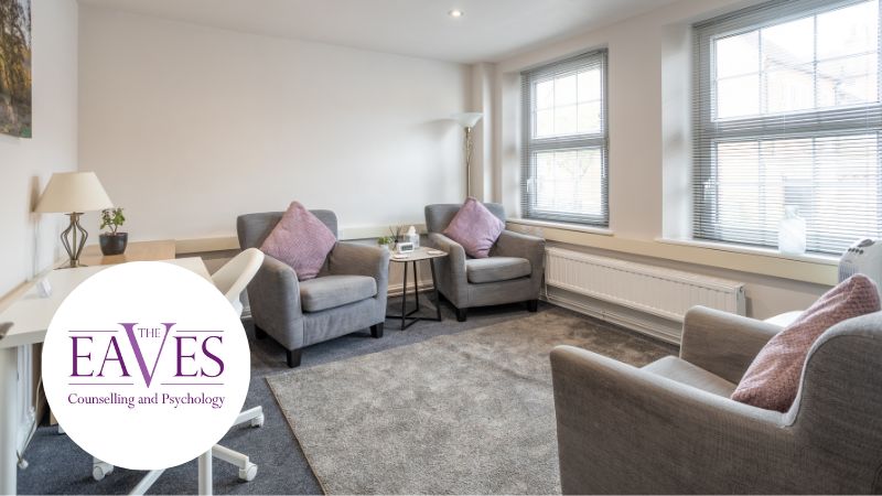 Modern therapy room with white desk, soft grey chairs, and neutral décor including a lamp, table, and tissues