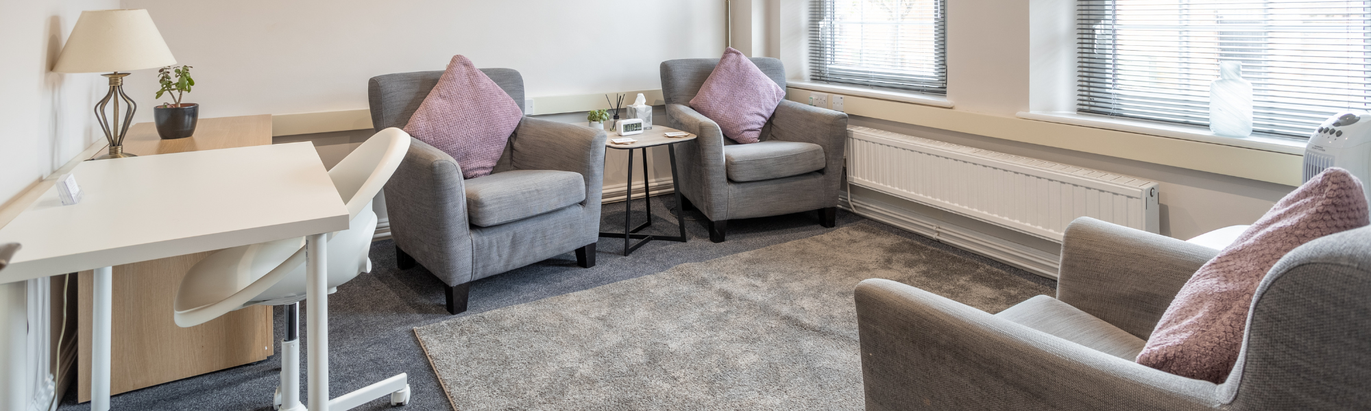 Modern therapy room with white desk, soft grey chairs, and neutral décor including a lamp, table, and tissues