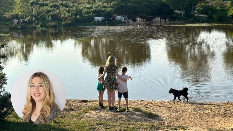 Three children standing by a lake with a dog approaching, siblings embracing while looking at water with trees and cows reflected in the background