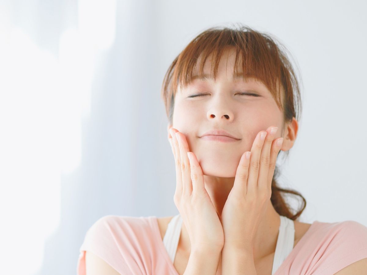 Woman with eyes closed gently smiling with hands on her cheeks against a soft neutral background, representing self-compassion and healthy self-esteem