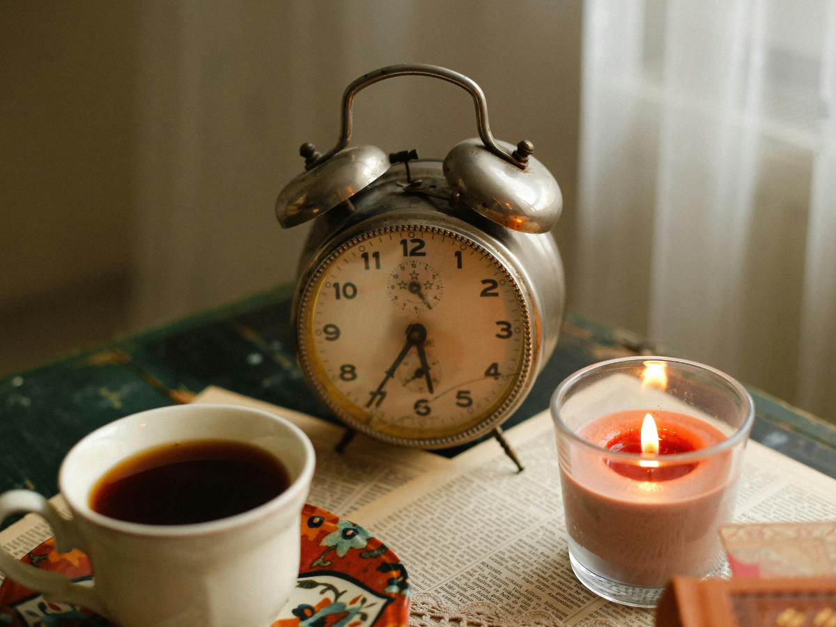 Close-up of an old-fashioned alarm clock, cup of tea and burning candle on a table near a window curtain, symbolising calm and reflection