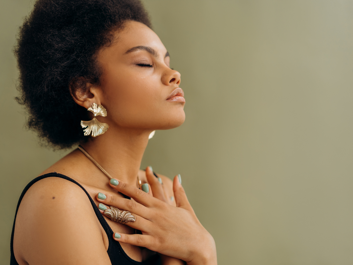 Black woman with afro hair, eyes closed, holding her hands across her chest while practicing a mindful breathing technique.
