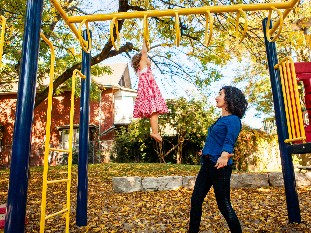 Mother watching her daughter play on climbing frame at playground, supporting confidence and active play for child development