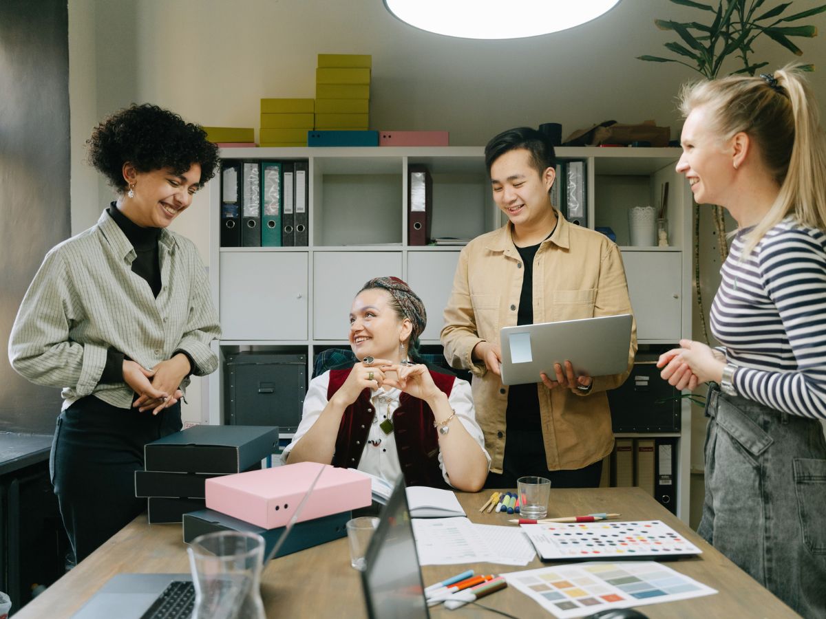 Employees smiling and collaborating around a table with laptops, papers and files, with a bookshelf in the background, highlighting workplace mental health support and team engagement.