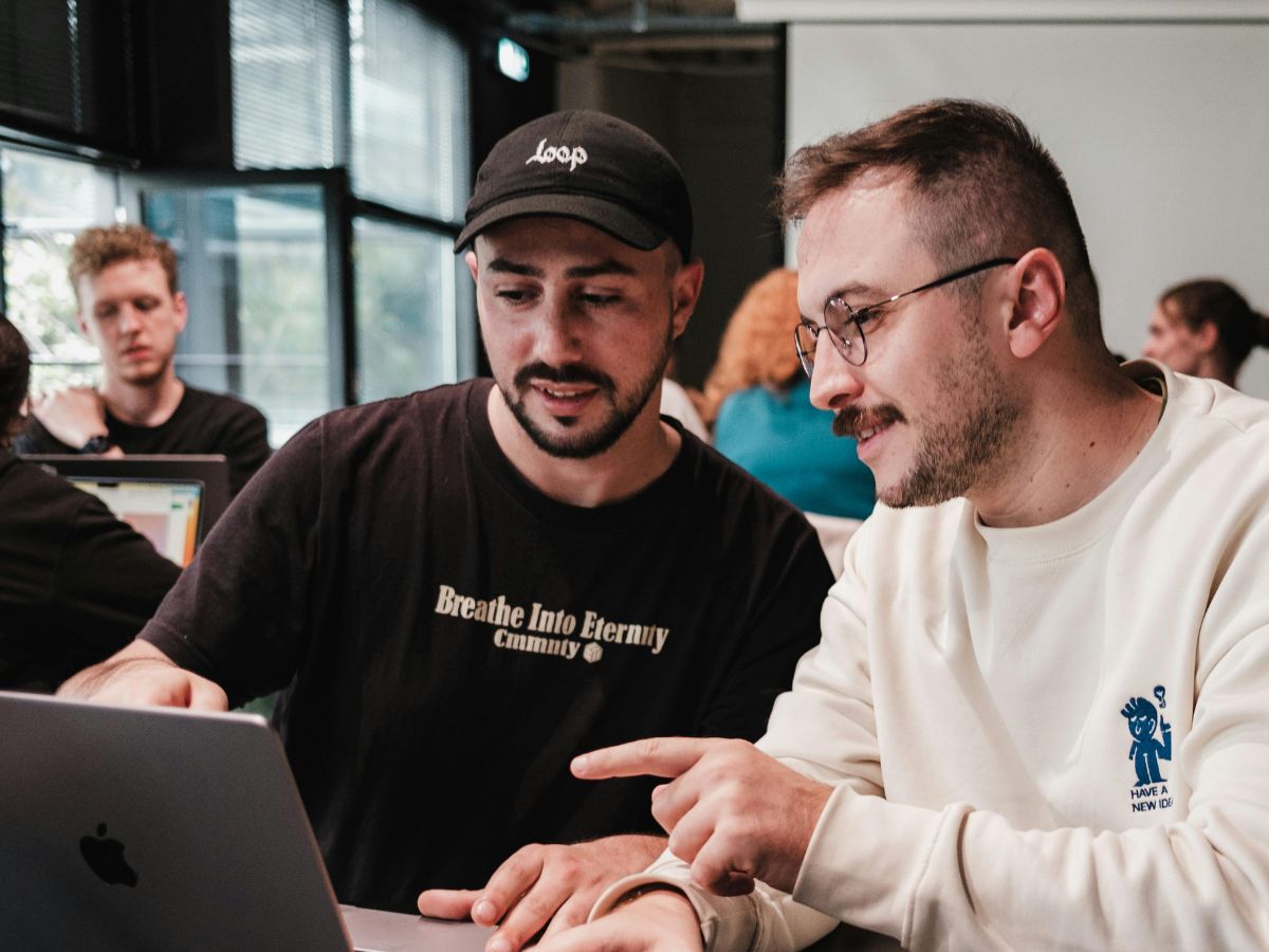 Two male employees discussing work in front of a laptop with colleagues blurred in the background, illustrating workplace mental health support and collaborative teamwork.