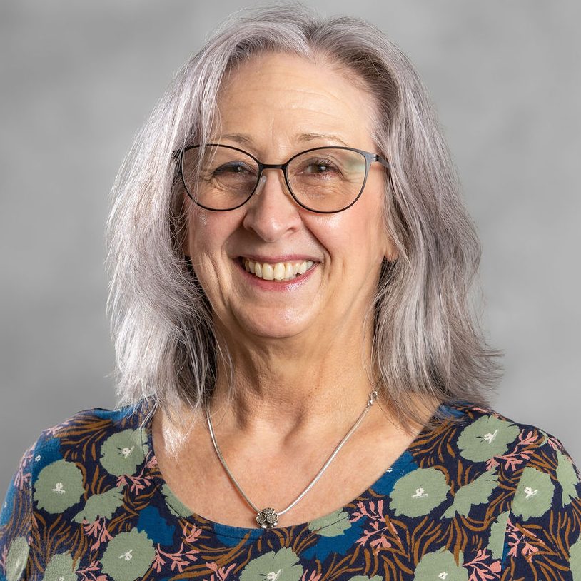 Image of Fiona Rimmer, an integrative psychotherapist working with adults experiencing difficult attachment and relational dynamics. She has long grey hair and is wearing a blue floral shirt with green and red patterns on.